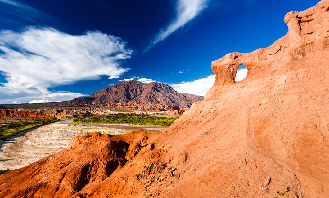 Red rock formations with a hole, a riverbed, green vegetation, and a distant mountain under a blue sky with scattered clouds in the Magic Triangle of the Andes desert landscape.
