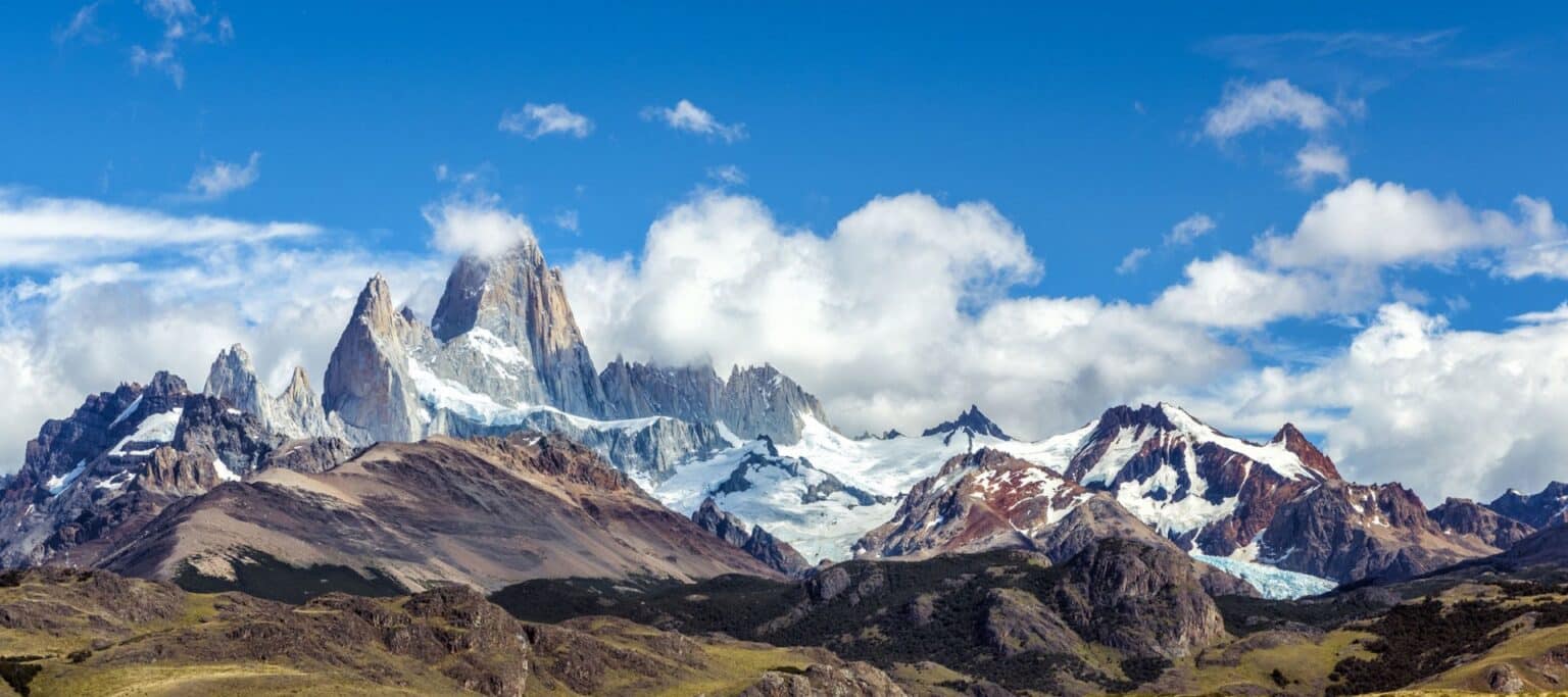 Snow-capped mountains with rugged peaks rise under a bright blue sky with scattered clouds, while rocky slopes and green grassy terrain extend in the foreground—an unforgettable sight on Argentina tours.