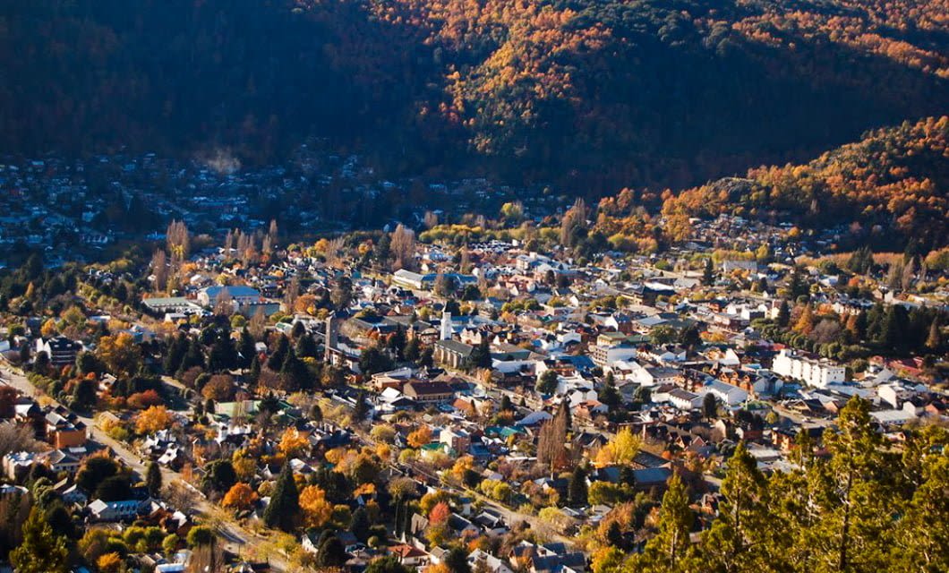 A small town surrounded by forested alpine hills with trees in autumn colors. The town has many houses, buildings, and streets nestled closely together under soft, natural light for a true Alpine experience.