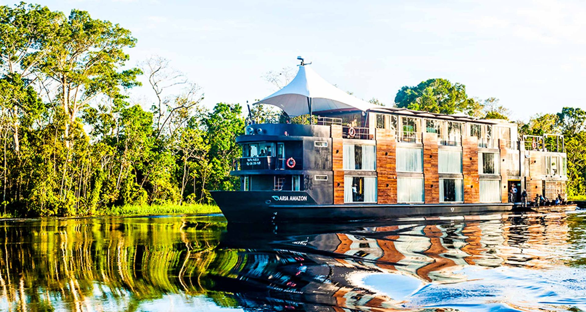 A modern river cruise ship with large windows and wooden paneling sails on a calm, reflective river in the Peruvian Amazon, surrounded by lush green trees under a clear sky—a true Aria Amazon Cruise luxury experience.