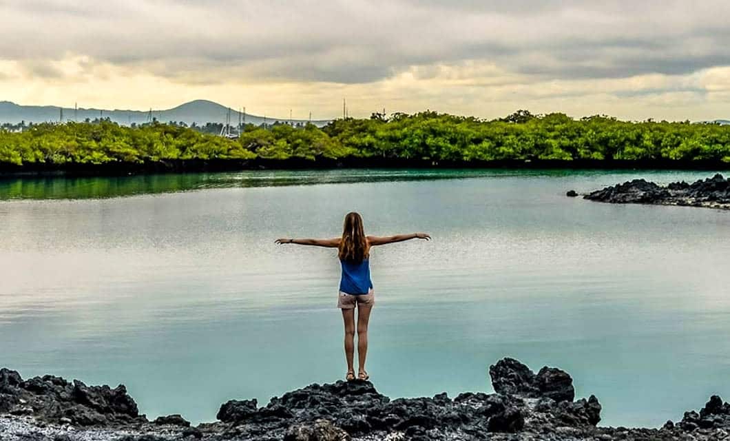 A person stands on black volcanic rocks with arms outstretched, facing a calm blue lagoon in the Galapagos, bordered by green trees and teeming with wildlife under a cloudy sky—a perfect moment on a Discovery Tour.