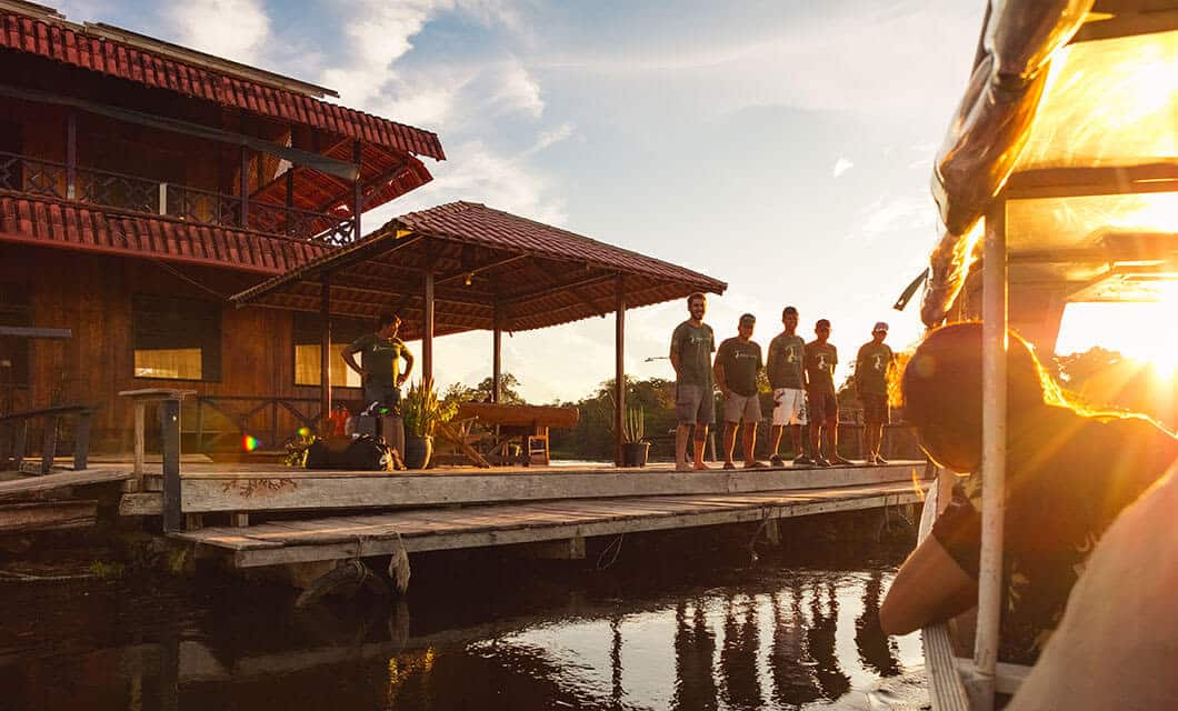 A group of people stand on a wooden dock by the water near a rustic lodge at sunset, enjoying the warm, peaceful atmosphere of a Remote Amazon Tour, while another person observes from a boat in the foreground.