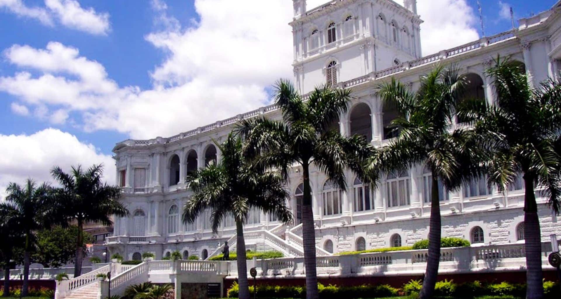 A grand white neoclassical building with arches and columns, partially obscured by tall palm trees in front, stands under a bright blue sky with scattered clouds in Asuncion.