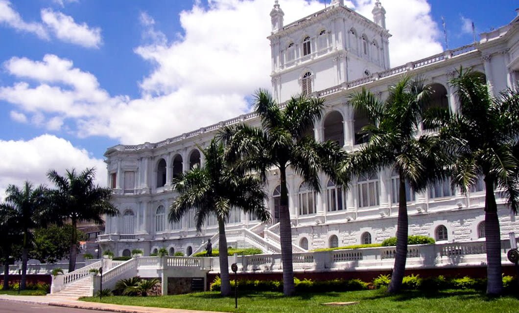 A grand white neoclassical building with arches, columns, and a central tower, surrounded by palm trees and green lawns under a partly cloudy blue sky—a must-see on any Paraguay Tour before heading to Iguazu.