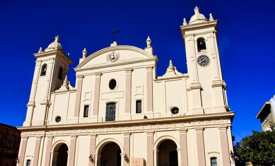 A cream-colored neoclassical cathedral with three arched entrances and twin bell towers stands proudly in Asuncion, set against a bright blue sky in the background.
