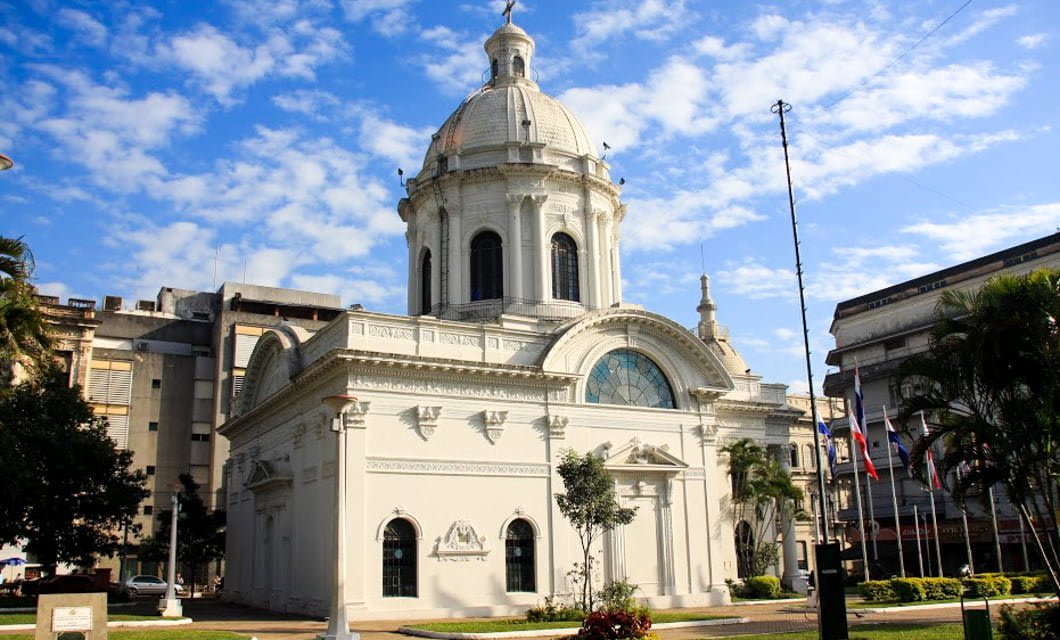 A grand white neoclassical building with a large dome and ornate details stands in Asuncion, surrounded by palm trees and flags, under a blue sky with scattered clouds.
