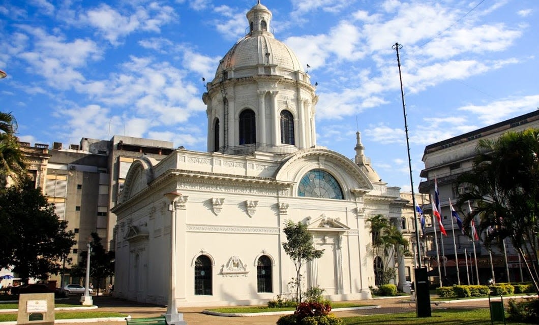 A historic white domed building with arched windows and ornate details stands under a blue sky, surrounded by a park where flagpoles display several national flags, evoking the rich heritage of the Incas.