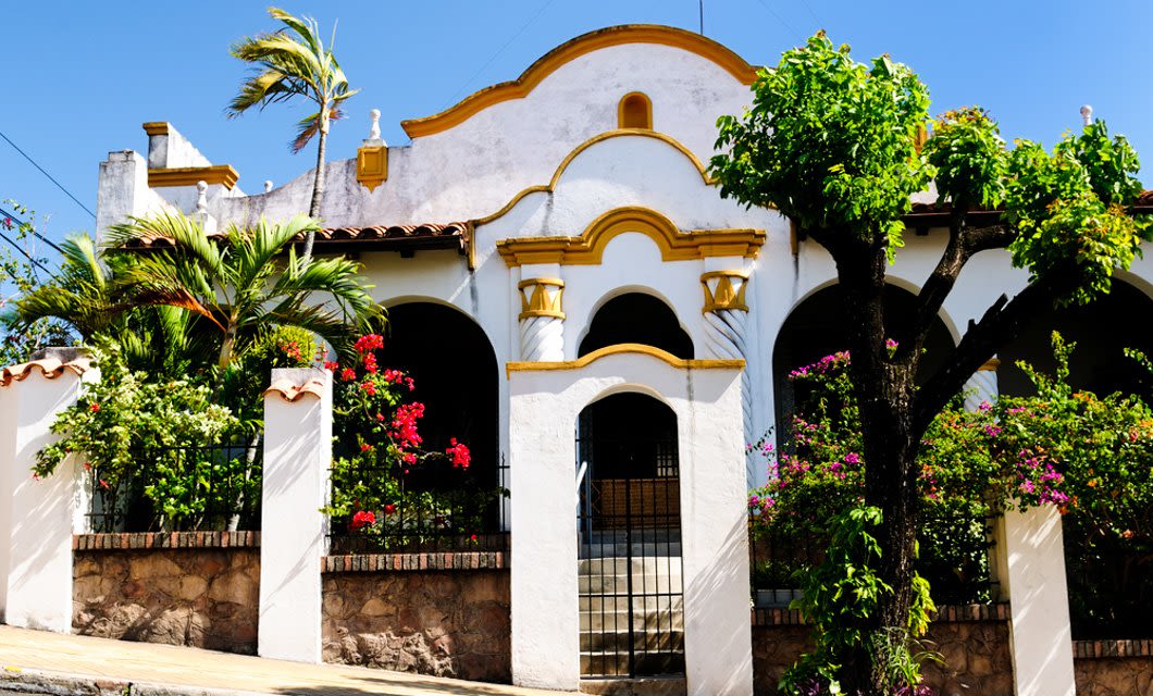 A white stucco house with yellow trim features arched doorways, decorative roof details, and is surrounded by flowering plants and palm trees, evoking the charm of Asuncion, with a stone fence under a clear blue sky.