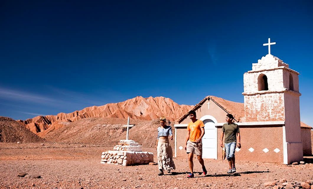 Three people walk on the dry, rocky Atacama Desert near a small white church with a bell tower and crosses. Reddish mountains rise behind them under Santiago’s deep blue sky.