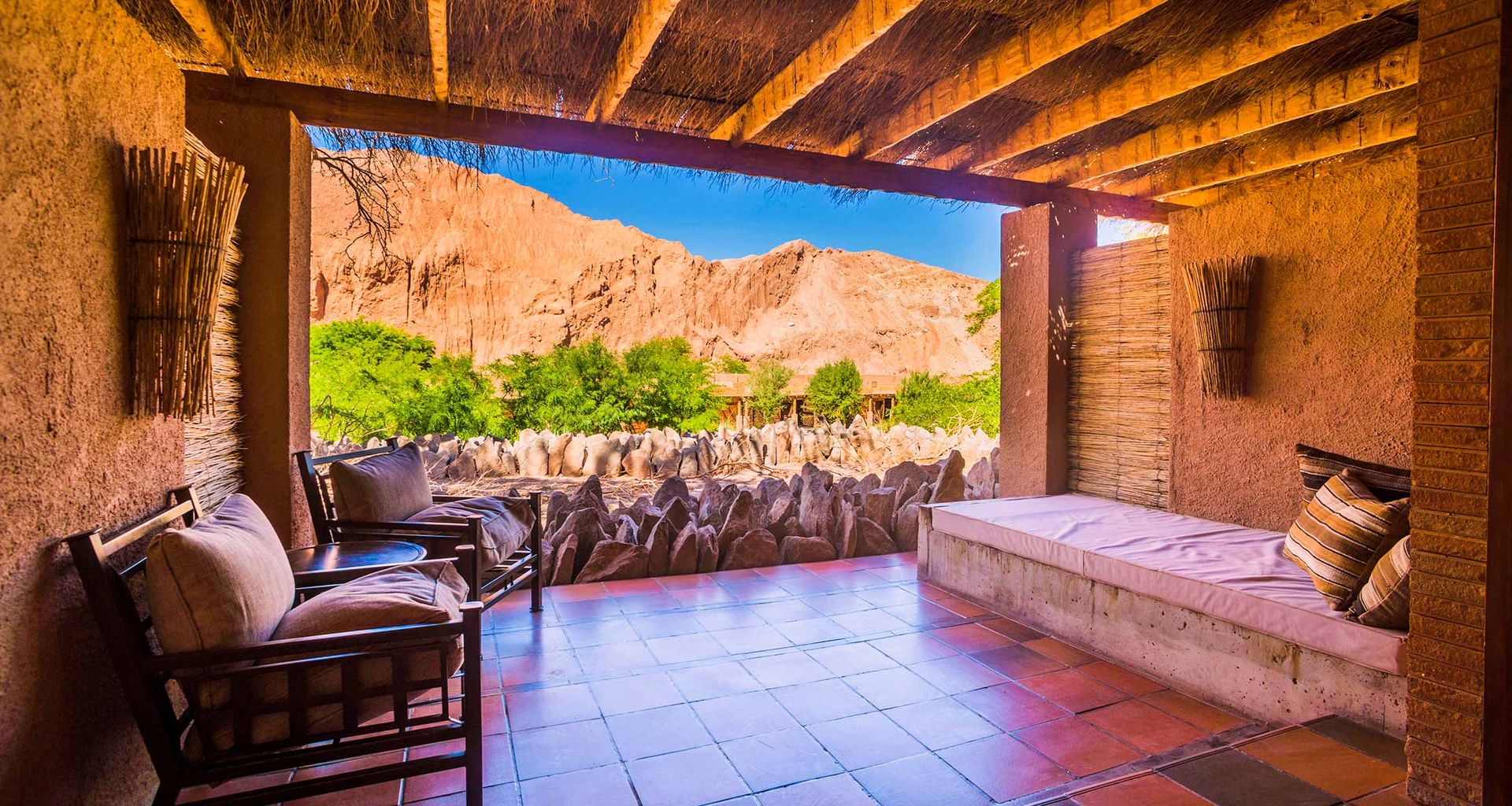 A shaded patio with cushioned seating at Alto Atacama overlooks a rocky landscape and distant mountains of the Atacama Desert under a bright blue sky, framed by wooden beams and rustic walls—a tranquil retreat in this luxury hotel.
