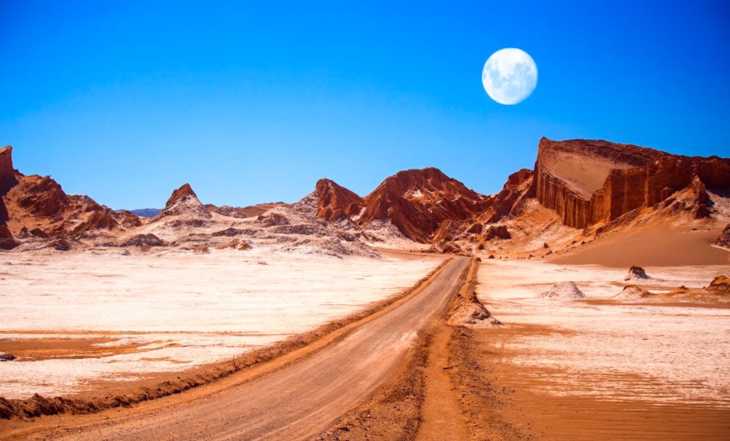 A sandy dirt road stretches through the Atacama Desert near Santiago, winding past orange rock formations under a bright blue sky. A full moon is visible above the horizon.