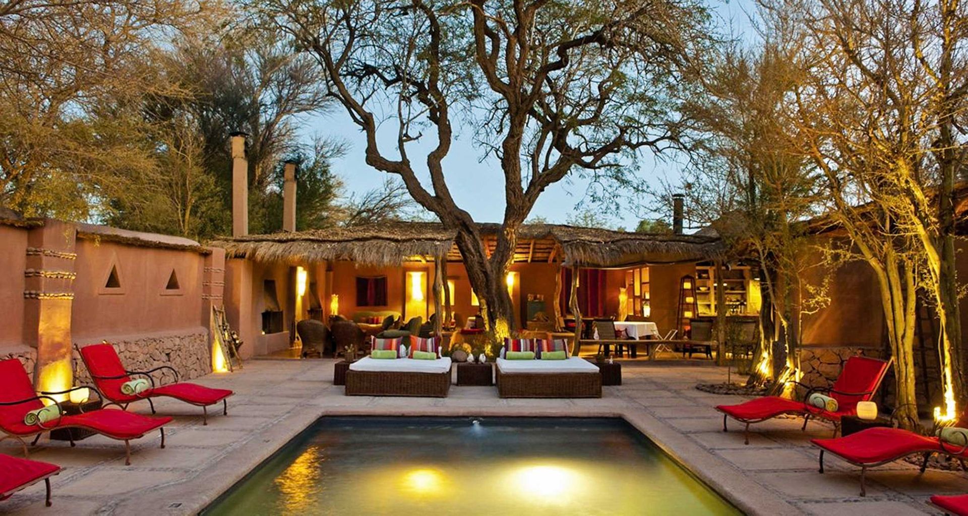 A cozy courtyard at Awasi Atacama features a small illuminated pool, surrounded by red cushioned lounge chairs, two daybeds, and a large tree. Warm lights and rustic adobe buildings create a serene outdoor setting in this luxury hotel in the Atacama Desert.