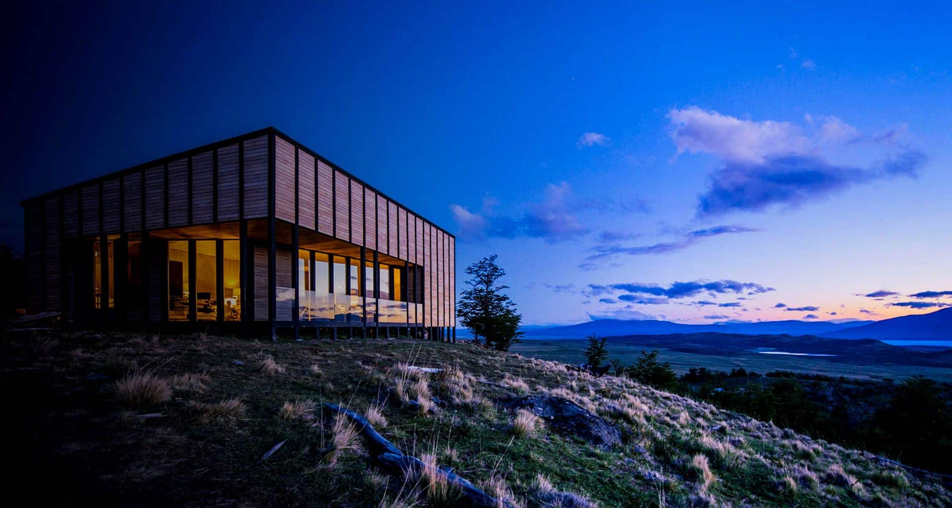 A modern wooden house with large glass windows sits on a grassy hill at dusk, overlooking a scenic landscape of rolling hills, water, and a blue and purple sunset sky—reminiscent of an exclusive luxury lodge in Torres del Paine.