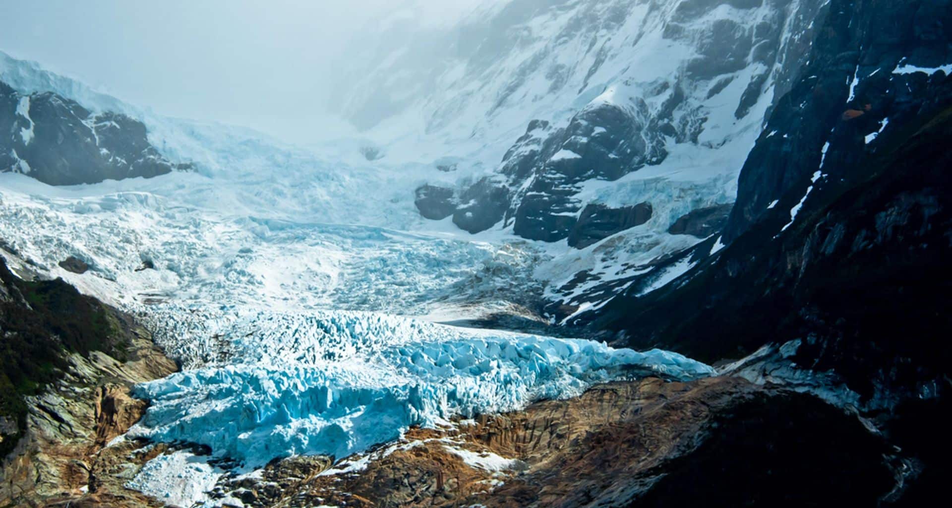 A stunning view of a glacier flowing down between rocky, snow-covered mountains—an iconic Patagonia sight—where bright blue ice contrasts against dark rock and patches of snow. Perfect for any Patagonia Essentials guided group tour adventure.