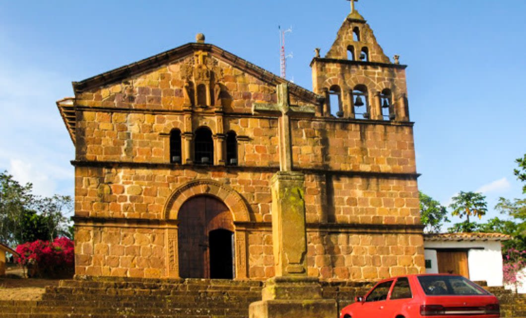 A historic stone church with a bell tower stands under a clear blue sky in Santander, Colombia. A red car is parked near the steps, and a tall stone cross marks the entrance—perfect scenery for your Colombia travel tour package.