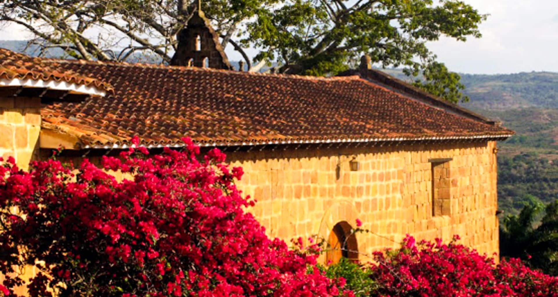 A rustic stone building with a red-tiled roof is bordered by vibrant red bougainvillea flowers, set against a backdrop of trees and distant hills under a bright sky—perfect for your next Colombia travel adventure.