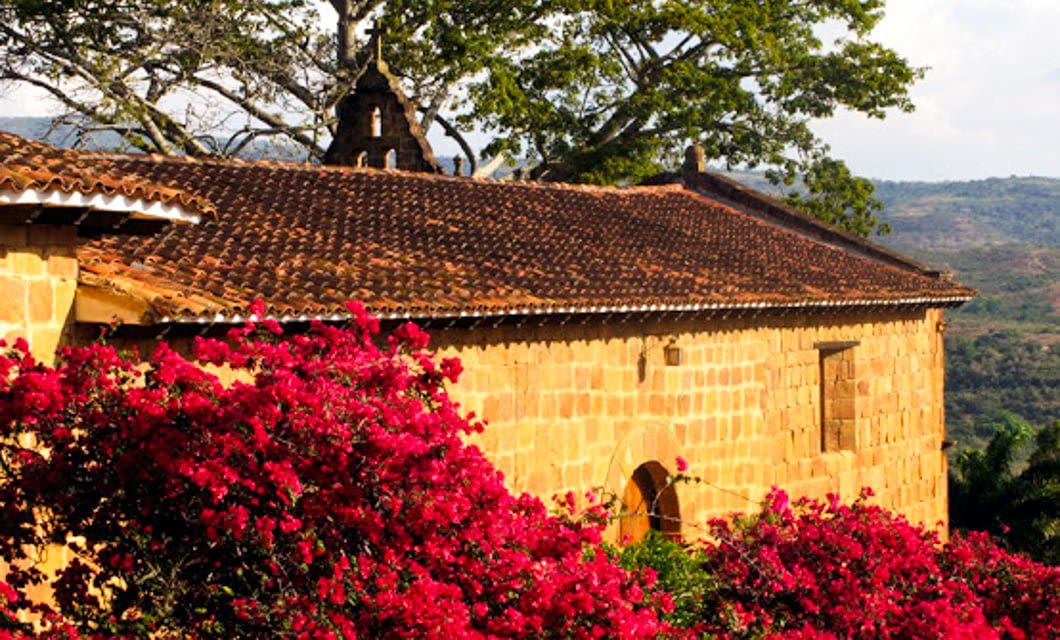 A rustic stone building with a tiled roof stands behind vibrant red flowering bushes, framed by trees and distant green hills—capturing the charm of Santander Colombia, perfect for any Santander Tour package under a clear sky.