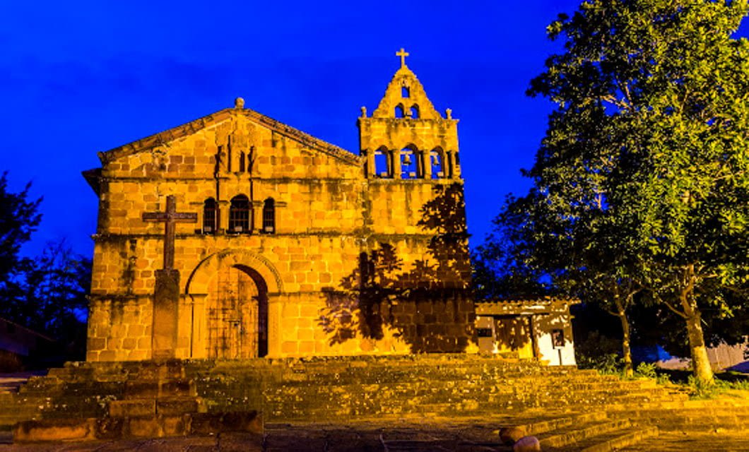 Stone church with arched doorway, bell tower, and cross at the entrance in Santander, Colombia, illuminated at night under a vivid blue sky with a large tree to the right—a stunning sight on your Colombia Travel tour package.