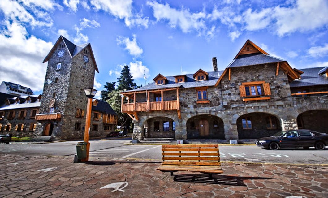 A stone building with wooden balconies and a tall clock tower stands under a partly cloudy Patagonia sky. A bench and streetlamp are in the foreground, with a black car parked nearby.