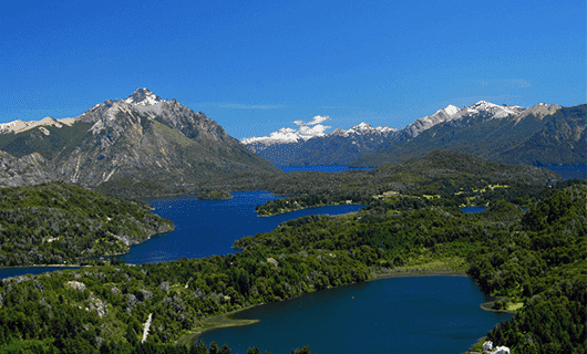 Patagonia mountain landscape with lush green forests, several blue lakes, and snow-capped peaks under a bright blue sky with a few white clouds—a perfect view from the scenic La Trochita train tour.