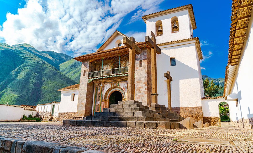 Colonial-era church with white walls, wooden balcony, and bell tower, set against green mountains near Cuzco and a blue sky, with stone crosses and a cobblestone plaza in the foreground.