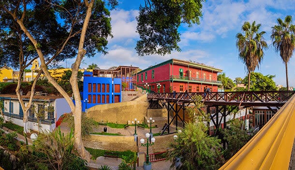Colorful buildings with red, blue, and yellow walls are surrounded by lush trees and greenery, evoking the lively spirit of South America's cultural capitals. A wooden bridge crosses a sunny courtyard under a bright blue sky.