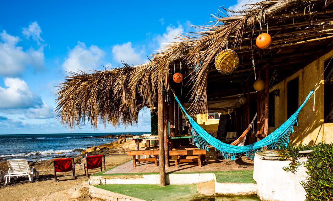 A rustic beach hut with a thatched roof features a blue hammock, wooden furniture, and hanging lanterns, overlooking the ocean in Piriapolis with chairs on the sandy shore under a bright, sunny sky.