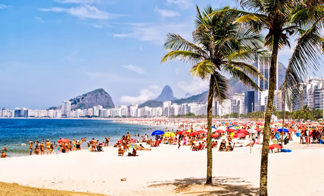Crowded sandy beach with colorful umbrellas, palm trees in the foreground, blue ocean, and a cityscape with tall buildings and mountains in the background under a sunny sky—perfect for your Brazil Tour experience.