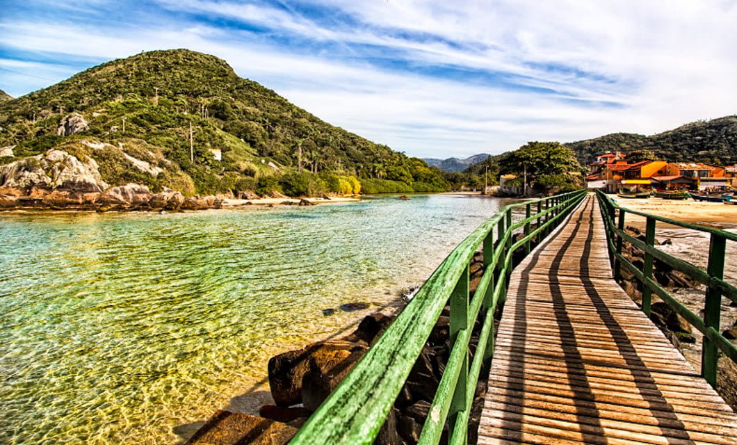 A wooden bridge with green railings stretches over clear, shallow water toward a sandy beach, offering a glimpse of adventure in Southern Brazil, with green hills and colorful houses under a partly cloudy sky in the background.