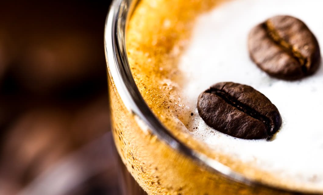 Close-up of a glass of frothy coffee topped with two whole coffee beans, showcasing creamy foam and bubbles—a true taste inspired by the rich Colonial Highlights of the Brazil Coffee Region.