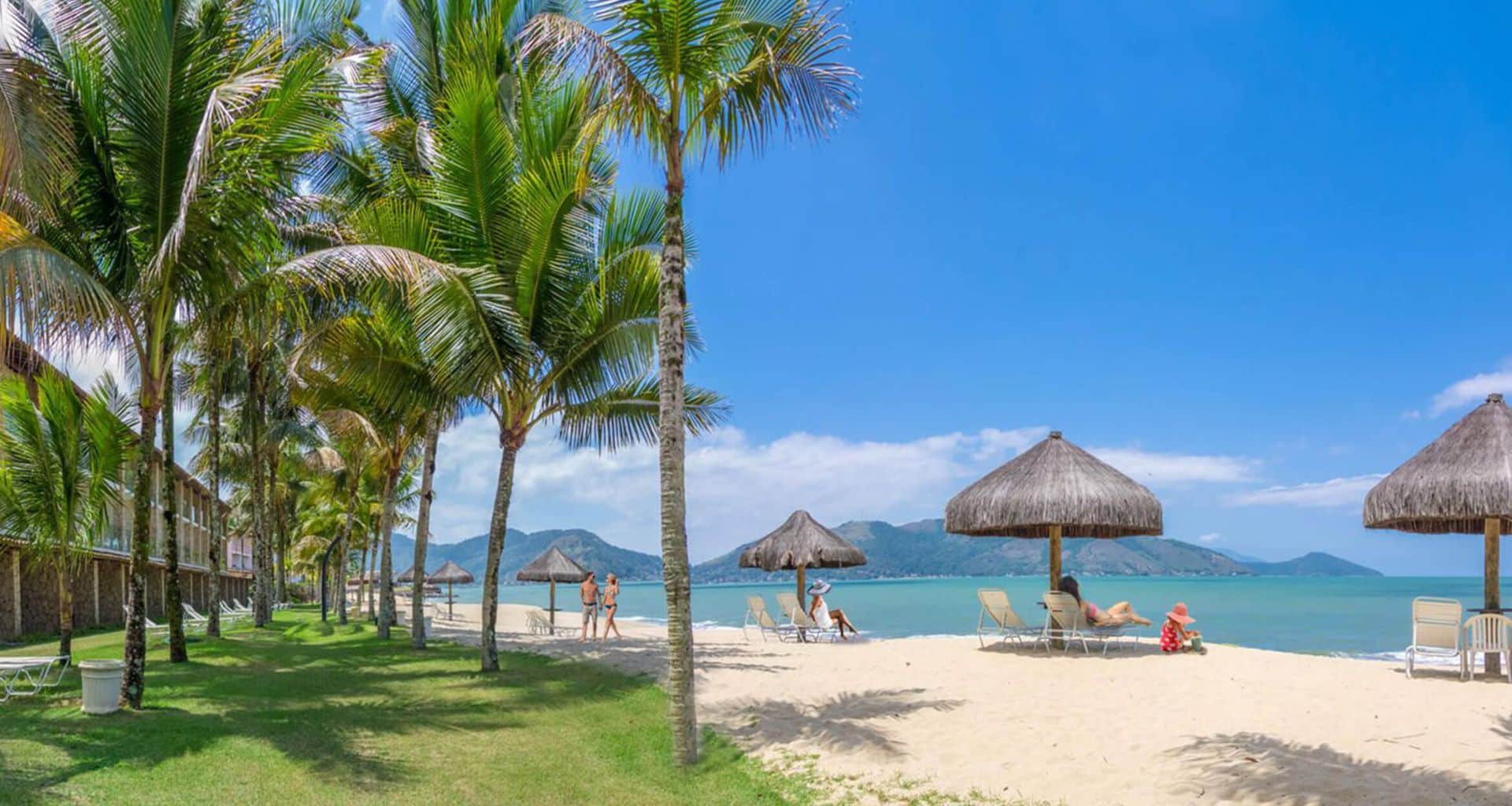 Palm trees line a sandy beach with straw umbrellas and lounge chairs at Portobello Hotel Resort. People relax and walk along the Angra dos Reis shore, with calm blue water and mountain views in the background under a bright, clear sky.