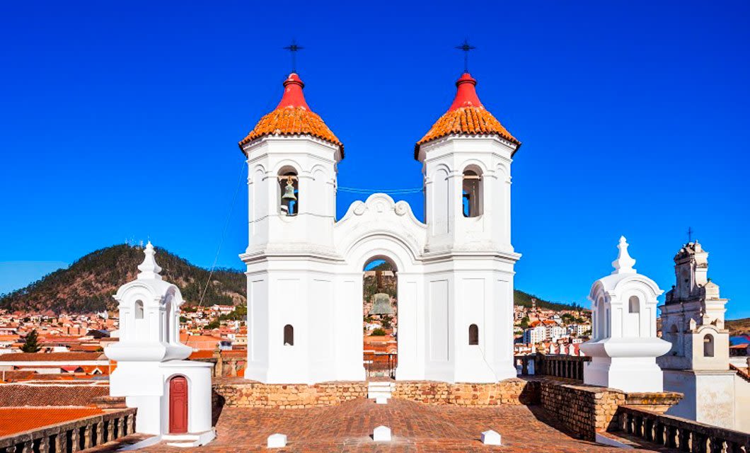 Twin white bell towers with red-tiled roofs stand on a rooftop overlooking a city of red-roofed buildings, with hills and a clear blue sky in the background—a classic scene among Bolivia highlights for any Bolivia travel adventure.