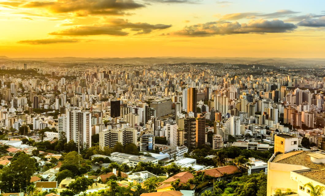 A panoramic view of a large city with numerous high-rise buildings and houses, set against a golden sunset sky with scattered clouds. The city, part of the Brazil Gold Route, is surrounded by distant hills.