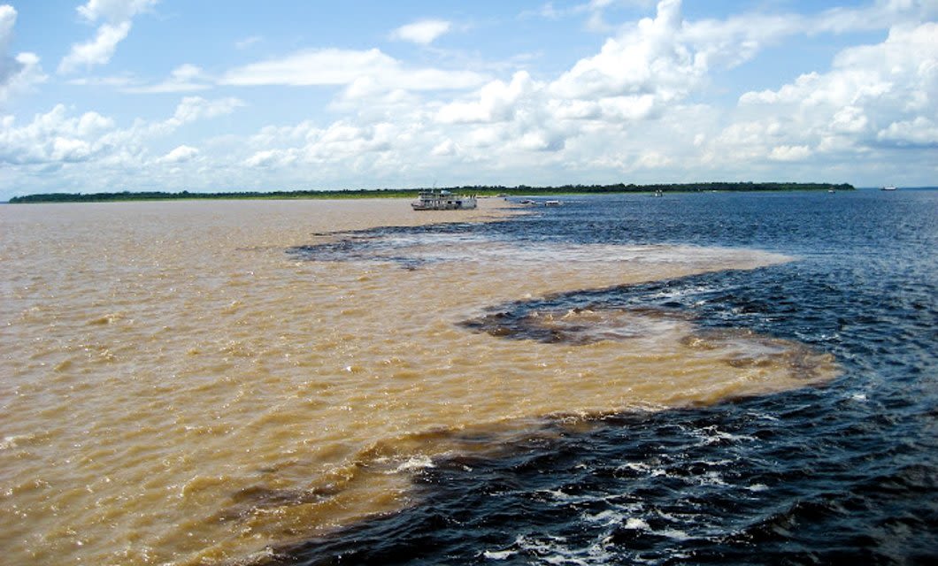 Two rivers with distinctly different colored waters, one brown and one dark blue, flow side by side without mixing under a partly cloudy sky. In the distance, a Tucano Amazon River Cruise boat drifts where the rivers meet.