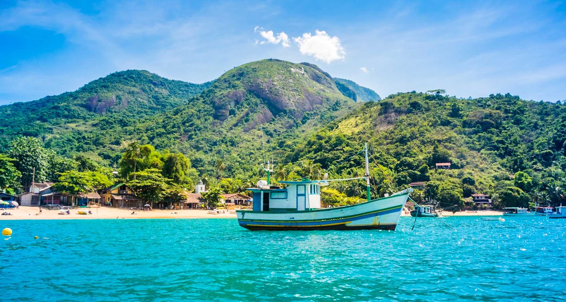 A colorful fishing boat floats on bright blue water near a tropical shoreline, with lush green mountains and scattered houses of Brazil’s colonial gems visible under a sunny, clear sky.