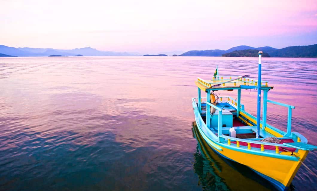 A brightly colored yellow and blue wooden boat floats on calm water at sunset, with pink and purple skies and distant hills evoking the charm of Brazil Coffee Region’s colonial highlights.