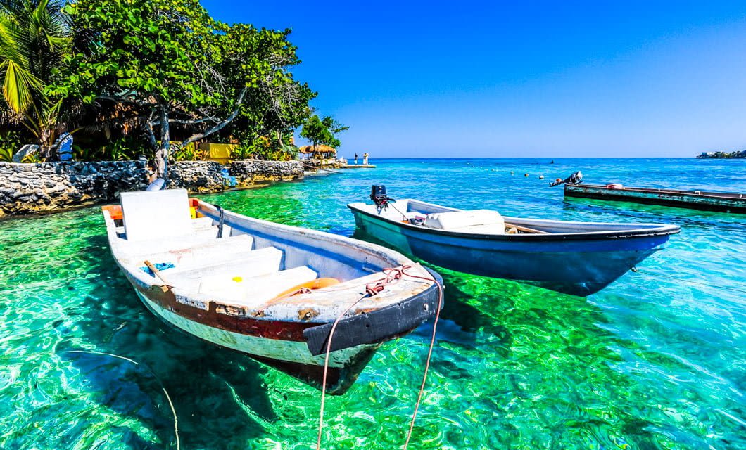 Two small boats float on clear turquoise water near a lush, green Colombian shoreline under a bright blue sky. The vibrant scene suggests a tropical, serene, and inviting coastal setting—perfect for a relaxing gourmet tour.
