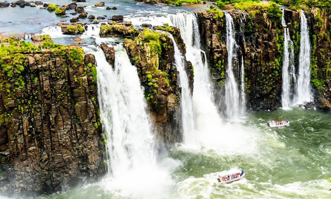 Several powerful waterfalls cascade down rocky cliffs surrounded by lush greenery. Below, three boats with tourists—many on a Brazil Train Tour—approach the base of the falls, creating white spray in the turbulent, greenish water.