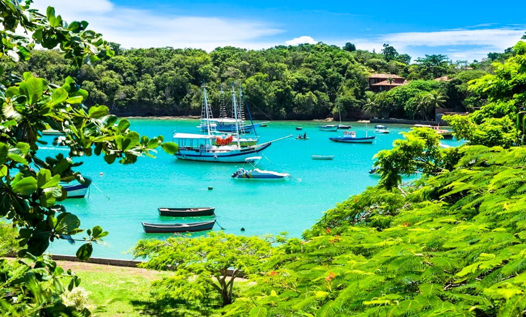 Bright blue tropical bay near Buzios Beaches with several boats and sailboats floating on calm water, surrounded by lush green foliage under a clear, sunny sky. Houses are partially visible in the background, capturing the charm of Rio de Janeiro.