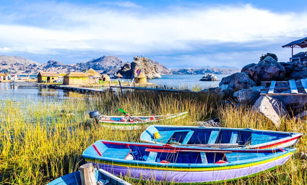 Colorful wooden boats rest among tall reeds on the shore of a lake, near floating Uros reed houses and distant mountains under a bright, partly cloudy sky.
