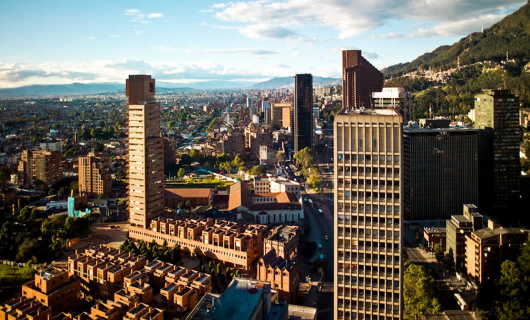 Aerial view of Bogota’s cityscape with tall modern buildings, tree-lined streets, and mountains in the background under a partly cloudy sky—an inviting scene for any Colombia tour.