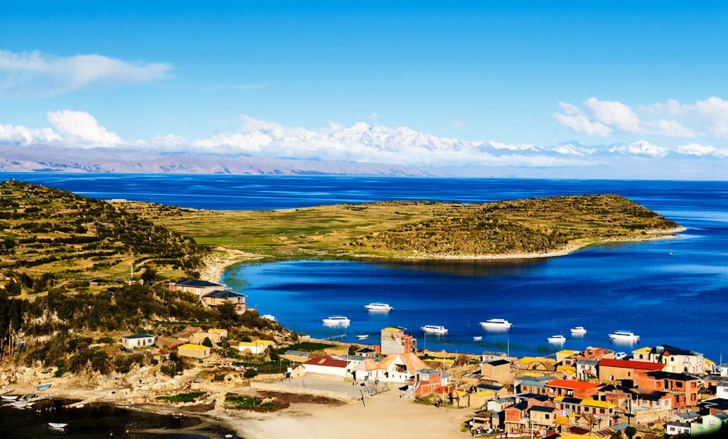 A coastal village with colorful houses sits beside a deep blue lake in Peru, with boats anchored on the water and green hills in the background. Snow-capped mountains and a bright blue sky complete this scenic landscape, perfect for a train tour.
