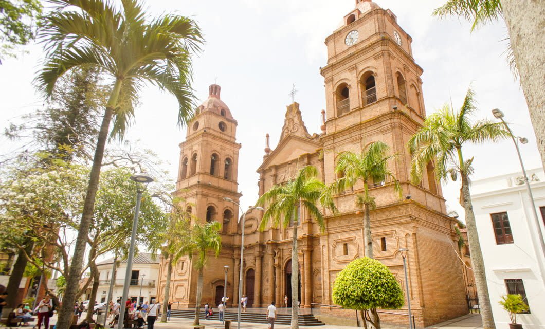 A historic brick cathedral with two towers stands among palm trees and people walking in a sunny plaza—an iconic stop on the Classic Bolivia Tour. The facade features arched doorways and a central clock above the entrance.