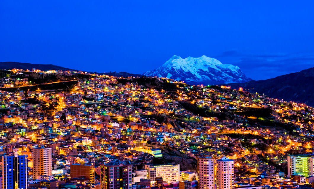 A vibrant cityscape at dusk with numerous glowing lights, nestled in hilly terrain—perfectly capturing the essence of a Classic Bolivia Tour—with a snow-capped mountain rising in the background under a deep blue sky.