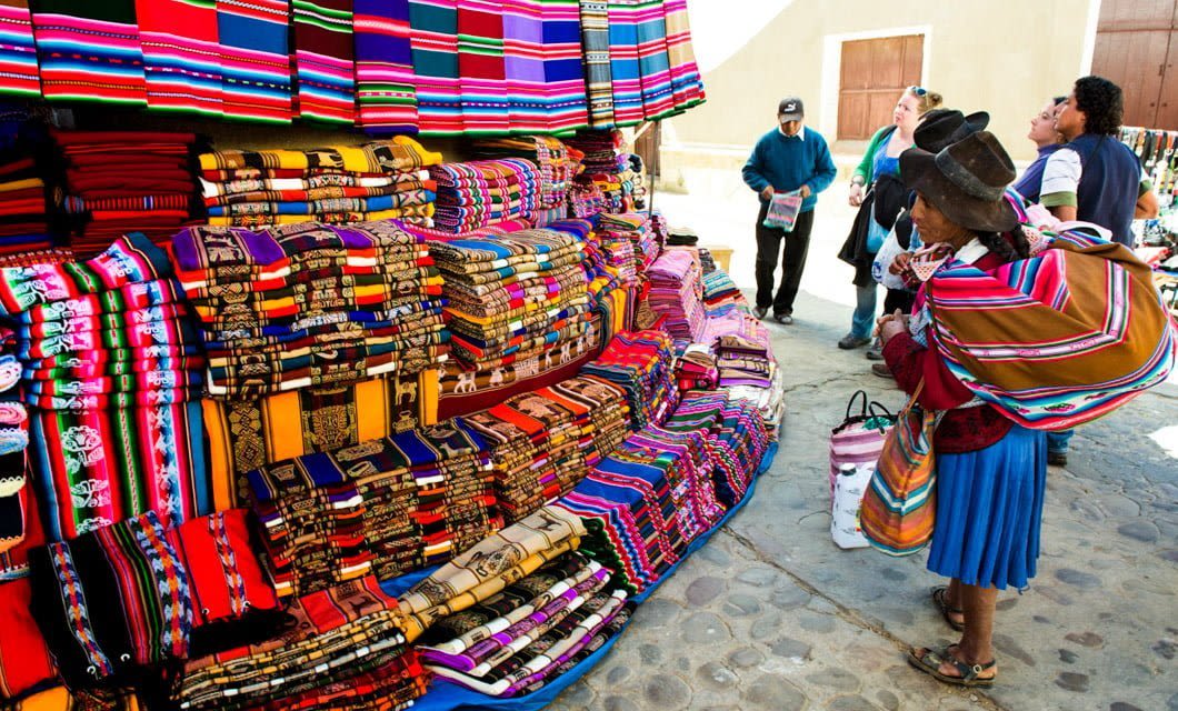 A woman in traditional clothing admires colorful textiles at a market stall outdoors, capturing the vibrant spirit of Bolivia travel, while other people stand nearby on a cobblestone street.