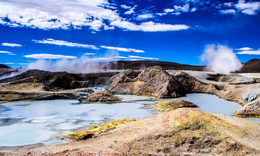 Geothermal area with steaming vents, rocky mounds, and bubbling pools under a bright blue sky—one of the Bolivia highlights to experience on any Bolivia tour. The barren landscape is dotted with yellow and white mineral deposits.