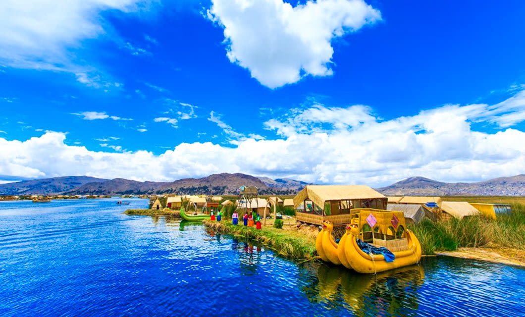 Colorful reed boats and houses line the edge of a blue lake under a bright sky, with mountains and grassy shores—an unforgettable sight for any Bolivia tour.