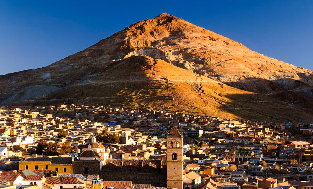 A densely packed town with colorful buildings sits at the base of a large, rugged mountain under a clear blue sky, bathed in warm sunlight—a scene straight from a Classic Bolivia Tour.