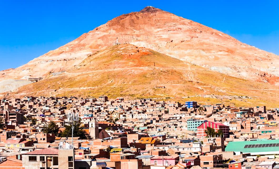 A sprawling city with brown-roofed buildings lies at the base of a large, reddish mountain under a clear blue sky—a stunning scene that captures the essence of Bolivia highlights. The mountain dominates, displaying layers of earthy colors.