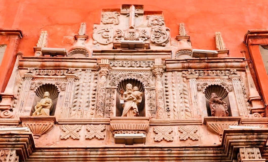 Ornate stone facade with three carved statues in niches against a red wall, featuring intricate architectural details, columns, and decorative shields above the statues—a striking example of Bolivia highlights for any Bolivia travel experience.